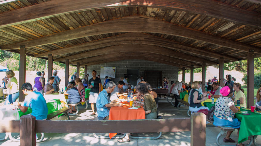 A photo of a group of people sitting under an outdoor gazebo, eating plates of food.