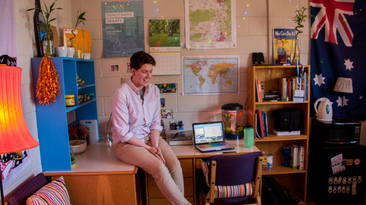 In the photo, a young woman (Maggie Carolan) sits on a desk in her dorm room. Her wall is covered with decorative posters, string lights, and a hanging Australian flag.