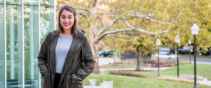 In the photo, a young woman (Erica Corder), wearing an olive coat over a gray shirt, stands and smiles for a posed photo in front of a courtyard with trees.