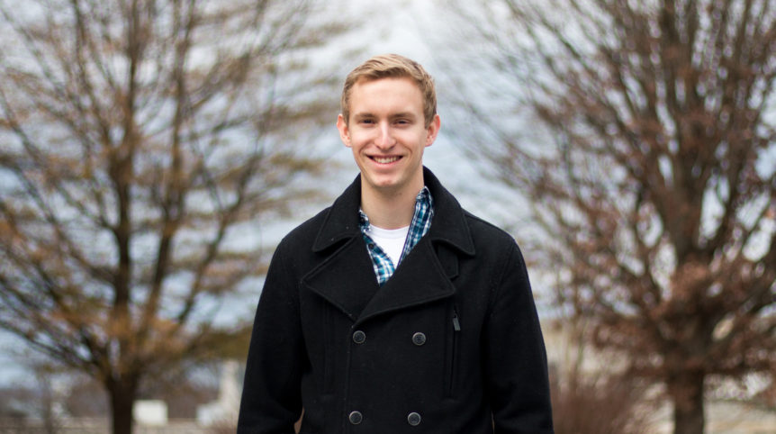 A white man wearing a black coat poses for a photo in front of a backdrop of trees.