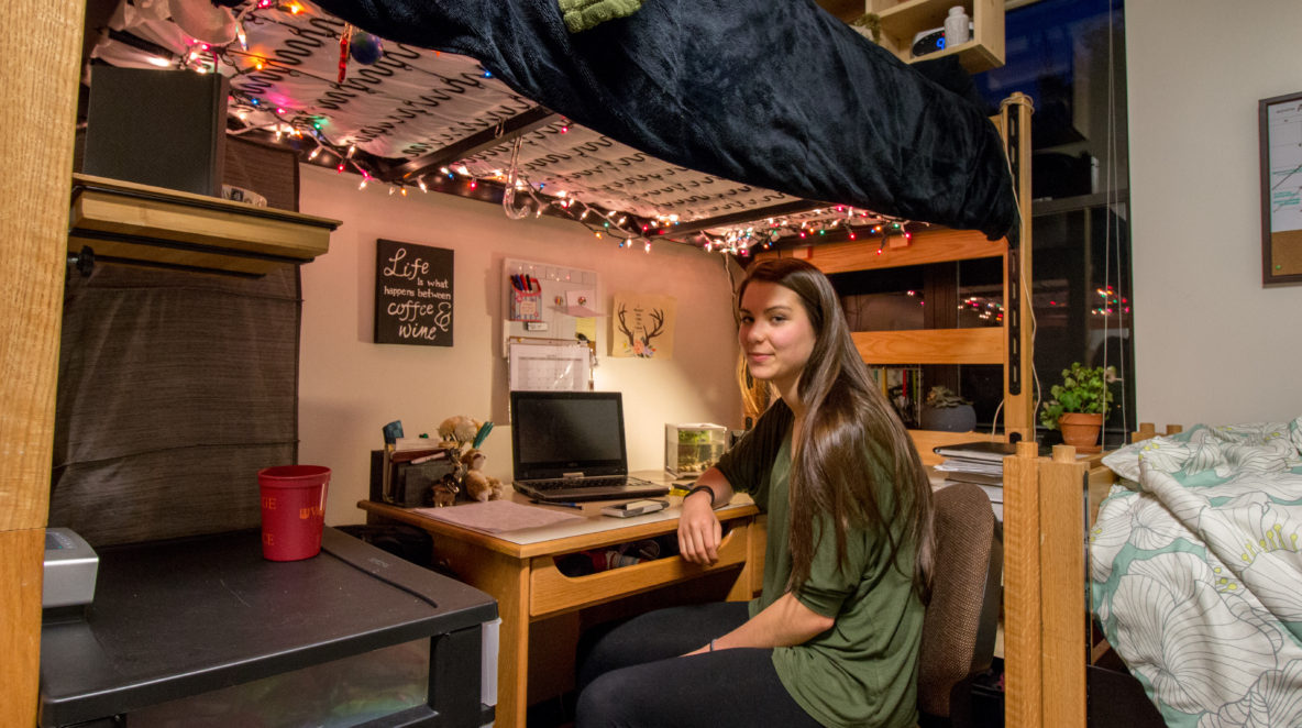 In the photo, a young woman (Emily Barritt) looks toward the camera for a portrait inside of her dorm room. She sits on a computer chair at a desk under a bunked bed, from which hangs multicolored string lights.