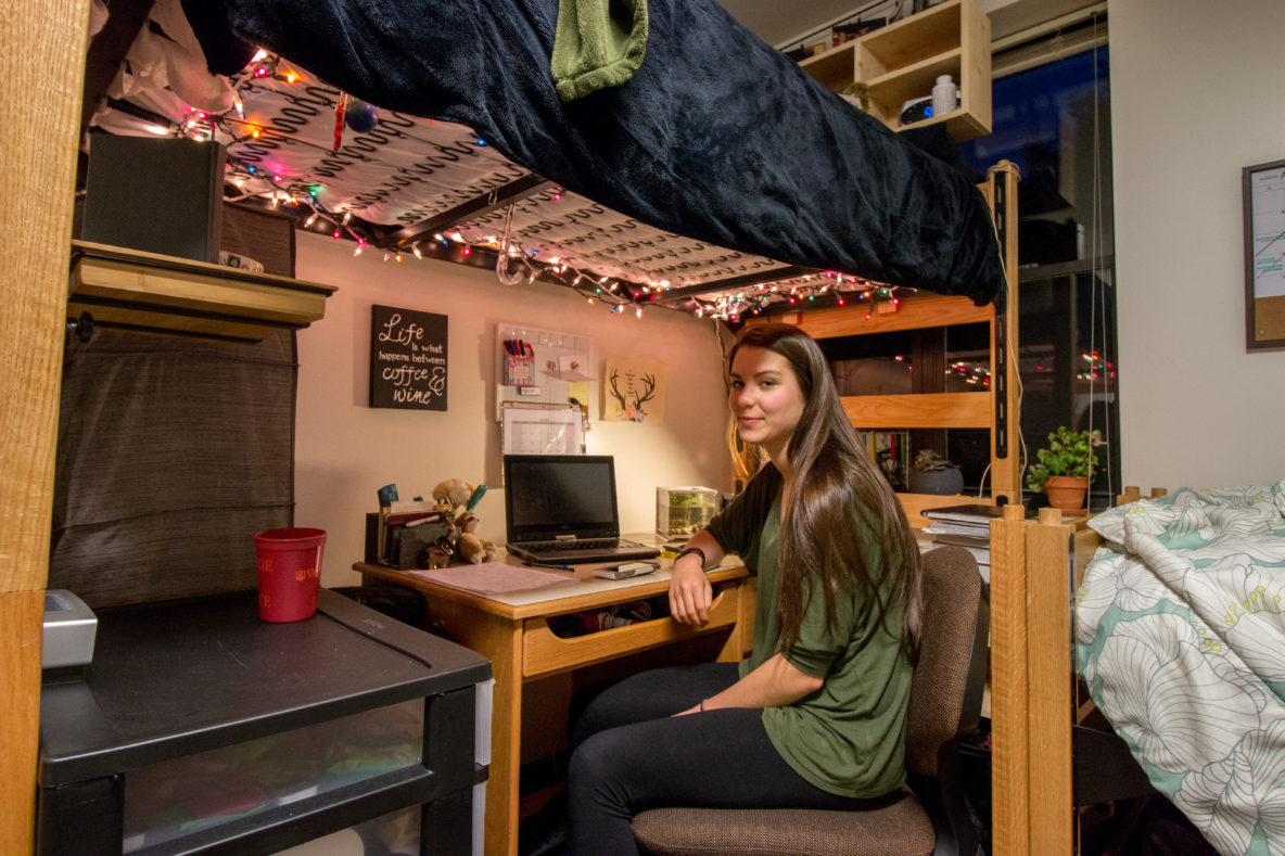 In the photo, a young woman (Emily Barritt) looks toward the camera for a portrait inside of her dorm room. She sits on a computer chair at a desk under a bunked bed, from which hangs multicolored string lights.
