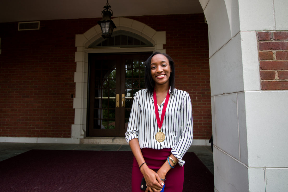 In the photo, a young Nigerian woman (Nneoma Nwankwo) stands with her hands clasped on the steps of a brick building. She is wearing a medallion that recognizes her as an Honors student.