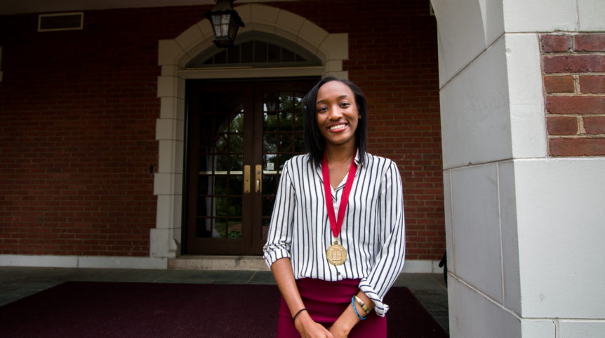 In the photo, a young Nigerian woman (Nneoma Nwankwo) stands with her hands clasped on the steps of a brick building. She is wearing a medallion that recognizes her as an Honors student.