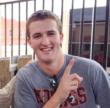 In the photo, a young man (Colin Moore) smiles and poses for a picture with one hand pointing up toward the sky to indicate number one. He is wearing a gray t-shirt with the word "Hokies" written in orange and maroon text.