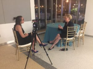 In the photo, Erica Corder sits next to a DSLR camera set up on a tripod that is recording an interview with a woman who is sitting across from her in a chair.