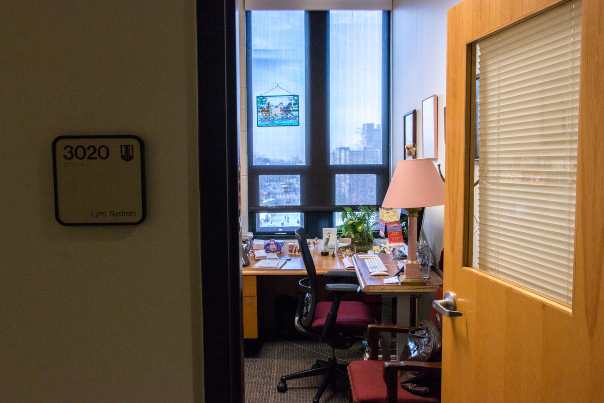 Photo taken from the outside of a small office. A sign with the name "Lynn Nystrom" is visible on the periphery of the photo. Inside the office is a desk and chair and an assortment of knick-knacks like photo frames and a stained glass picture of a horse.