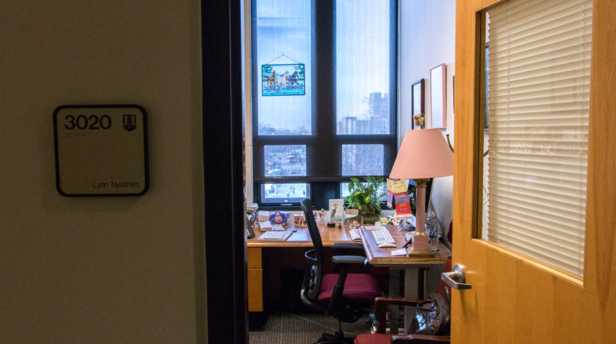 Photo taken from the outside of a small office. A sign with the name "Lynn Nystrom" is visible on the periphery of the photo. Inside the office is a desk and chair and an assortment of knick-knacks like photo frames and a stained glass picture of a horse.