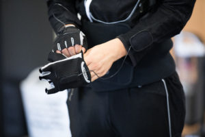 In the photo, a young man tries on a black cloth glove, which is connected to wires. It's a close up shot, focused only on the lower torso and the black suit.