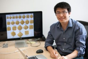 In the photo, a young man, Hongliang Xin, sits at a computer desk with an illustration of polymers pulled up on the computer screen behind him as he smiles toward the camera.