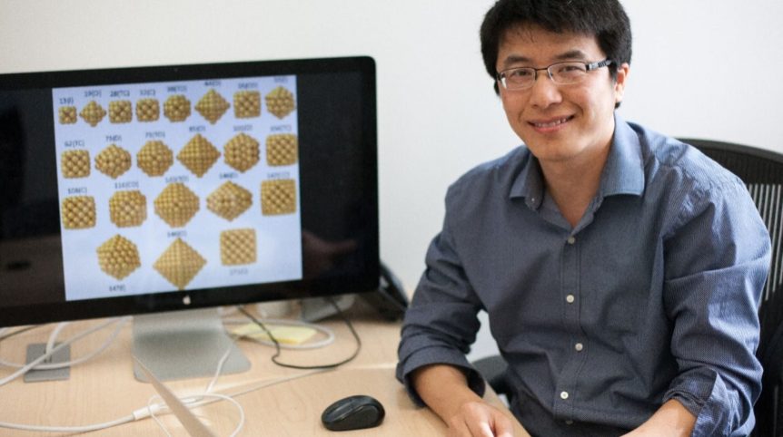 In the photo, a young man, Hongliang Xin, sits at a computer desk with an illustration of polymers pulled up on the computer screen behind him as he smiles toward the camera.