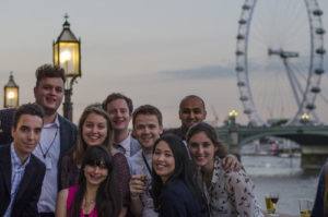 A photo of a group of smiling people standing in front of the London Eye at dusk.