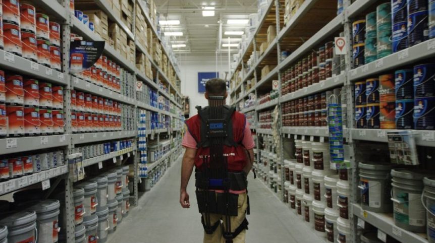 A man wears a soft robotic suit through an isle in Lowe's, surrounded by paint cans.