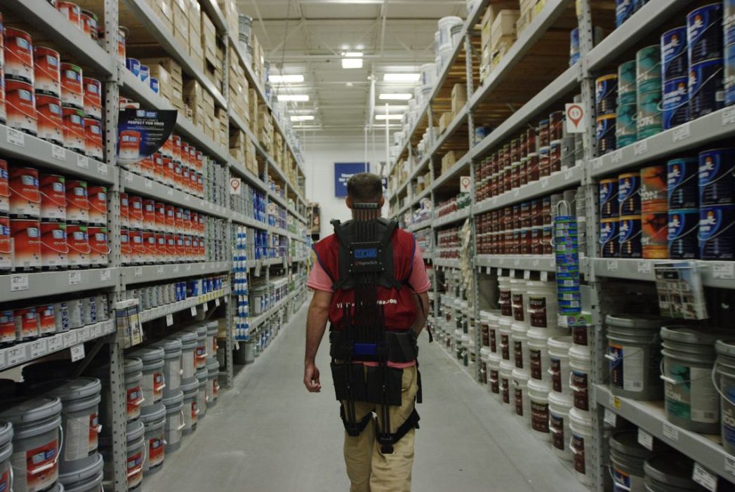 A man wears a soft robotic suit through an isle in Lowe's, surrounded by paint cans.