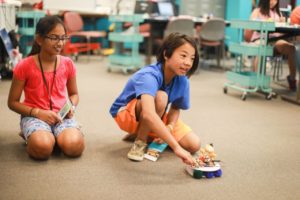 Two young girls sit on the floor with a robot they created that moves across the room.
