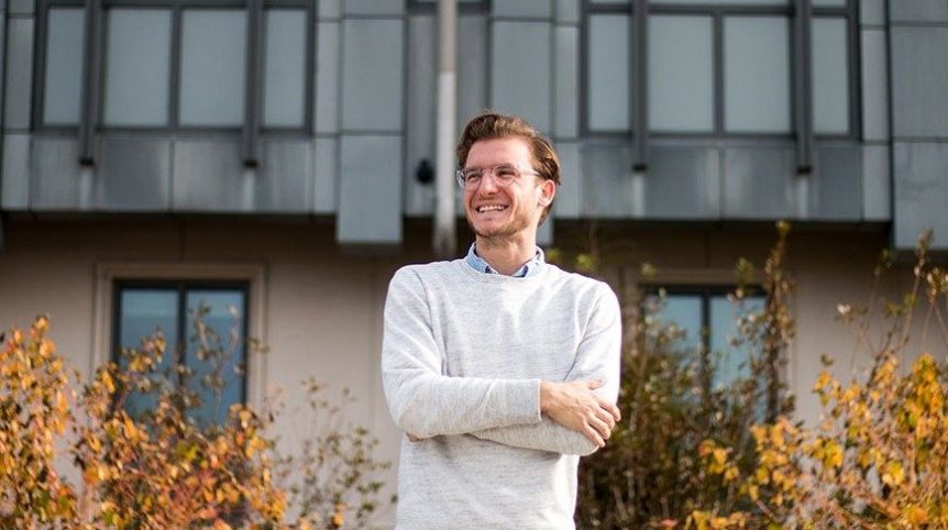 A posed photo of a young man in glasses standing in front of a building and fall foliage.