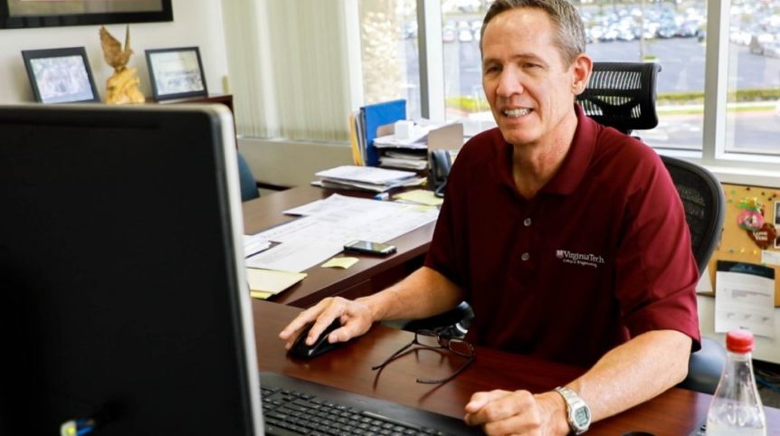 A man sits at a computer and works, wearing a maroon polo shirt that bears a logo from the Virginia Tech College of Engineering.