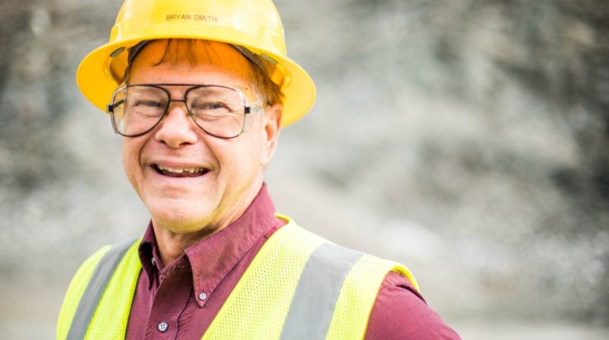 A man wearing a yellow hard hat flashes a smile for a photo taken inside a rock quarry.