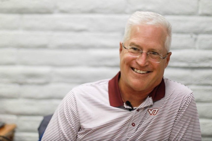 A man wearing a maroon-striped polo smiles for a photo inside a white-brick-walled office.