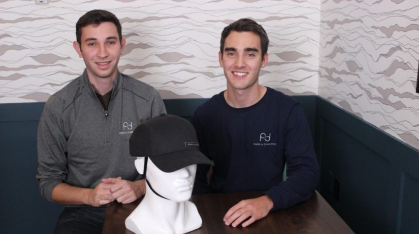 Two young men sit at a table and pose for a photo. On the table is a white mannequin head and neck, wearing a black helmet that looks like a baseball cap.