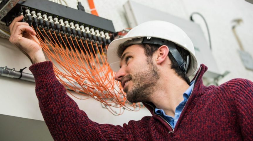 A man wearing a white hard hat and a red sweater reaches toward the ceiling, where he adjusts orange wires linked to a data acquisition system.