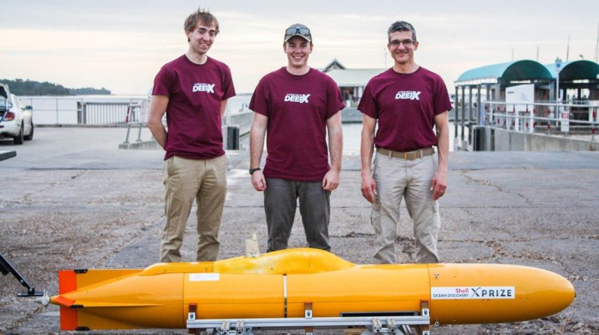 Three men stand behind a small yellow underwater autonomous vehicle and pose with smiles for a photo.