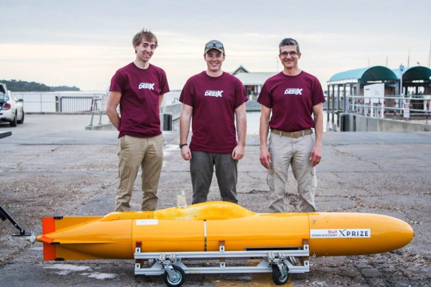 Three men stand behind a small yellow underwater autonomous vehicle and pose with smiles for a photo.