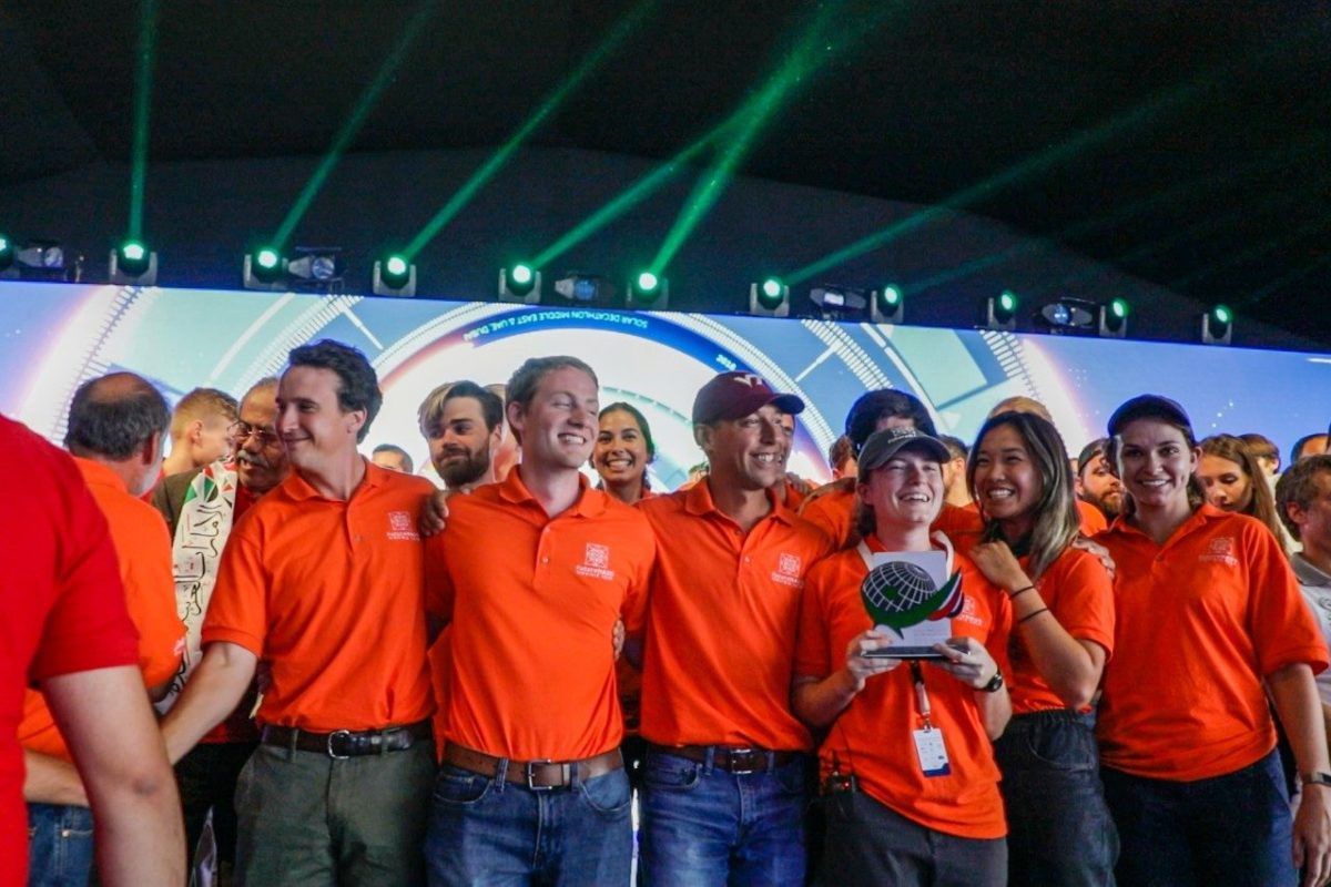 Students in orange team polos smile on a stage while one woman holds a trophy.