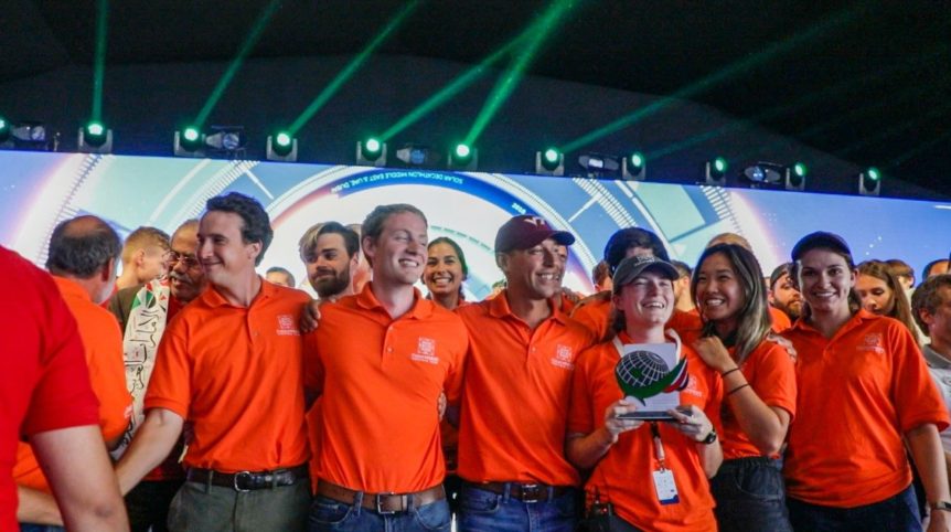 Students in orange team polos smile on a stage while one woman holds a trophy.