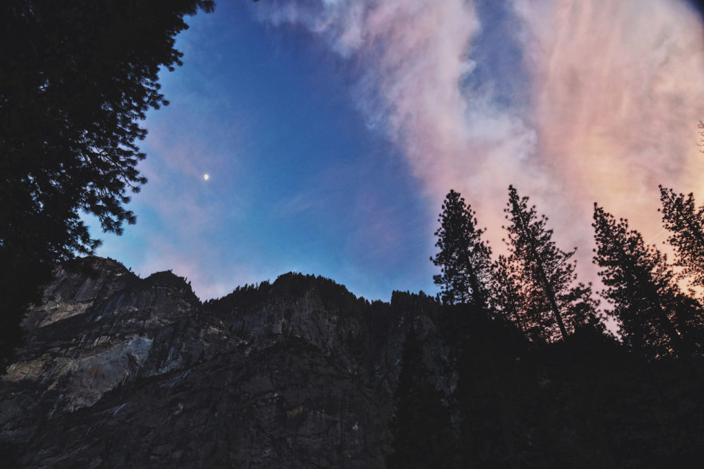 A photo of an evening sky, painted pinks and blues, with a moon shining over a treeline and mountainside.