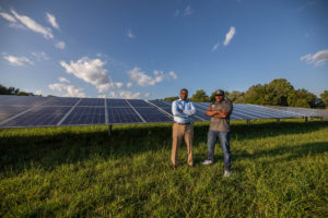 A photo of two men standing with their arms crossed in front of solar panels.