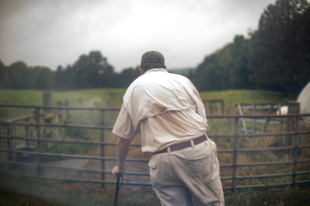 A photo of a farmer leaning on a cane in front of a gate.