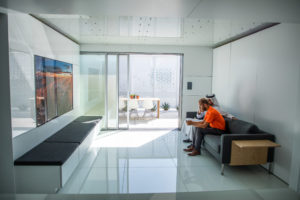 A photo of a white, futuristic home. Inside, two men sit on a couch and chat.