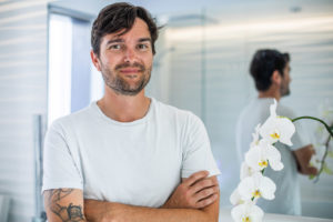 A photo of a young man with scruff, crossing his arms, wearing a white shirt, and smiling for a photo in front of a mirror background.