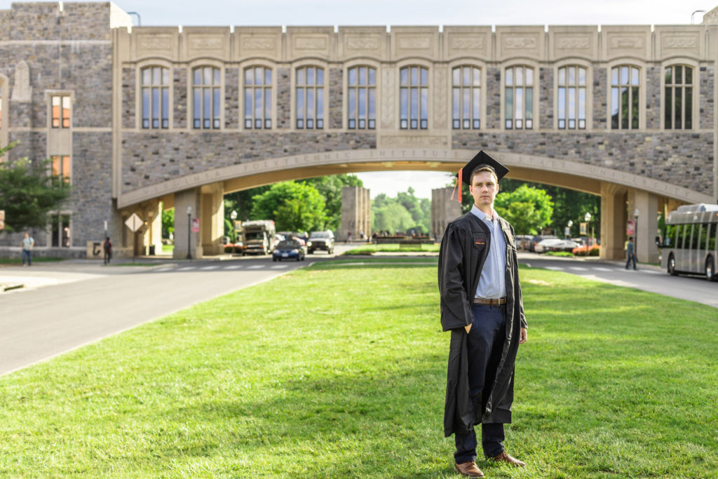 A photo of a graduate wearing a cap and gown and standing in front of an iconic bridge on the Virginia Tech campus.