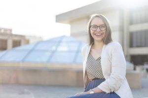 A woman poses for a photo, smiling. She's wearing glasses and sitting in front of a pyramid structure behind her.