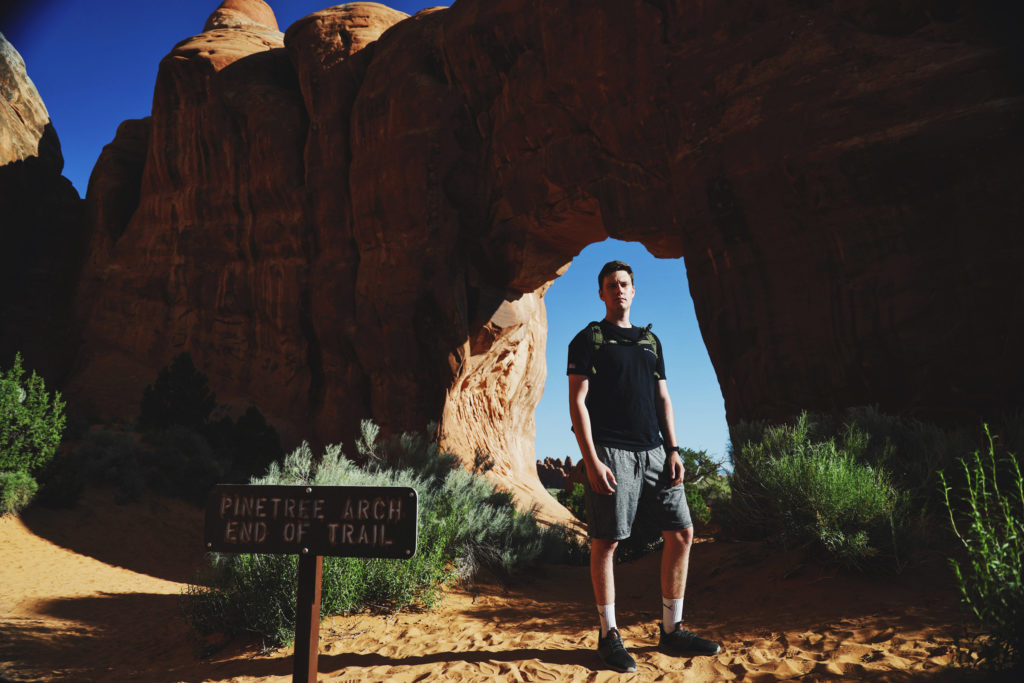 A photo of a young man standing in front of an arch. A sign in front of him reads "Pinetree Arch. End of trail."